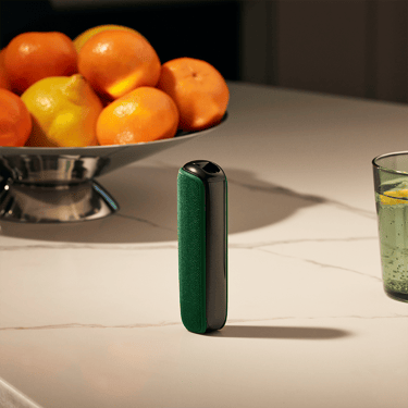 Ploom AURA ierīce ar Amazon Green aizmugurējo vāciņu uz galda blakus augļu grozam